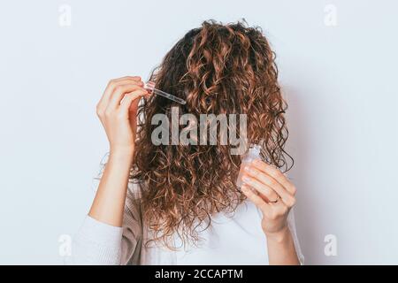 Donna che applica l'olio cosmetico ai suoi bei capelli ricci, primo piano su sfondo bianco Foto Stock
