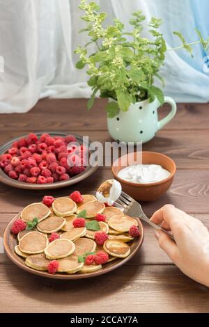 Pancake cereali in mano su una forchetta con yogurt. Lamponi freschi maturi rossi in un piatto marrone e un mazzo di Melissa in una tazza sulla linguetta di legno scuro Foto Stock