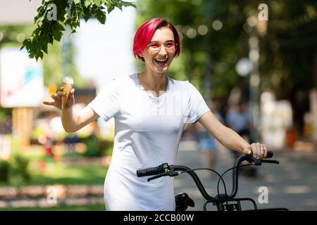 giovane donna con i capelli rosa cammina nel parco con una bicicletta che mangia gelato in estate. Modalità di trasporto ambientale Foto Stock
