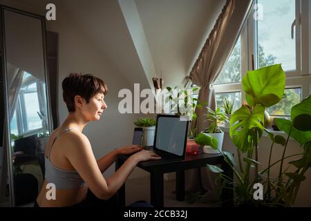 Donna di razza mista in abiti casual a casa lavorando su un computer portatile seduto ad un tavolo da caffè con molte piante verdi in vaso vicino alla finestra. Lavorare a casa Foto Stock