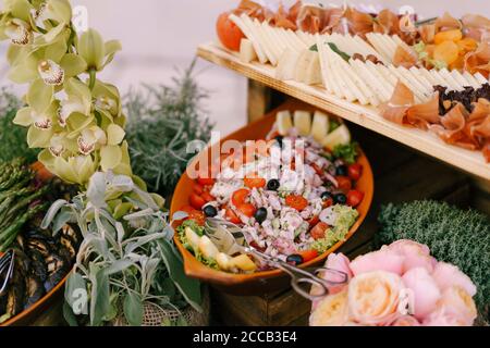 Insalata con pomodoro e olive nere in un piatto e affettati di formaggio e prosciutto su un tavolo con fiori. Foto Stock