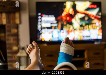 Il maschio sedette sul divano guardando la tv con un getto a piedi dalla quinta chirurgia metatarsale. Foto Stock