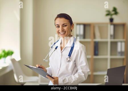 Giovane terapeuta sorridente medico che guarda la macchina fotografica e prende appunti sopra l'ufficio della clinica medica sullo sfondo Foto Stock