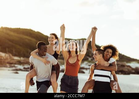 Amici multietnici di umore divertente godendosi sulla spiaggia. Uomini e donne allegri che si divertono a cavalcare con i piccini. Foto Stock