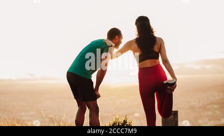 Fitness uomo e donna facendo esercizi di riscaldamento. Vista posteriore di un atleta femminile che allunga i muscoli in piedi all'aperto con il suo compagno di corsa. Foto Stock