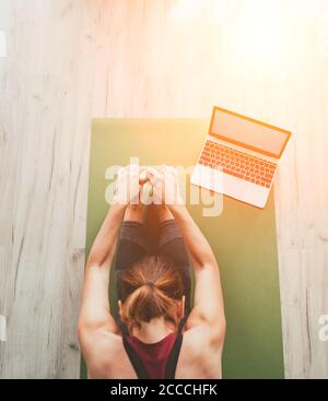 Donna sana e sportiva seduta sul tappetino in posa Paschimottanasana, facendo esercizi di respirazione, guardando una lezione di yoga online sul computer portatile. Sano p Foto Stock