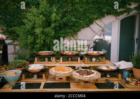 Cucina con padelle e tavole di legno per servire la pizza con cibo preparato per un banchetto sullo sfondo di un edificio con spazi verdi. Foto Stock