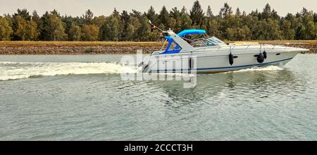 Gifhorn, Germania, 9 agosto 2020: Motoscafo bianco con tetto blu si muove ad alta velocità attraverso il canale con spruzzi d'acqua Foto Stock