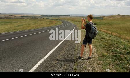 Un giovane scatta le immagini su un panorama del telefono seguente verso una strada di campagna Foto Stock