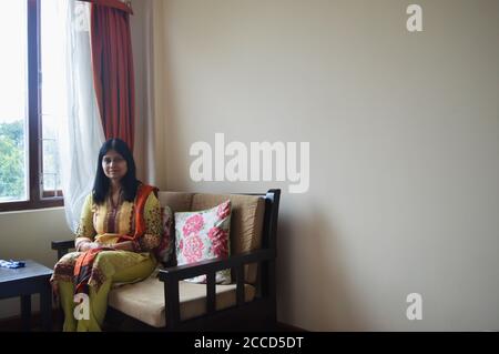 Una signora indiana seduta su un divano di legno vicino a una grande finestra di vetro, messa a fuoco selettiva Foto Stock