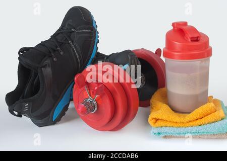 Concetto di stile di vita fitness. Manubri rossi, asciugamani colorati, shaker con proteine di siero di cioccolato e scarpe sportive nere. Oggetti di bodybuilding isolati Foto Stock