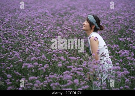 Una bella giovane donna asiatica in piedi in viola vervain Bush, sorridente felicemente. Sfocare sfondo Foto Stock