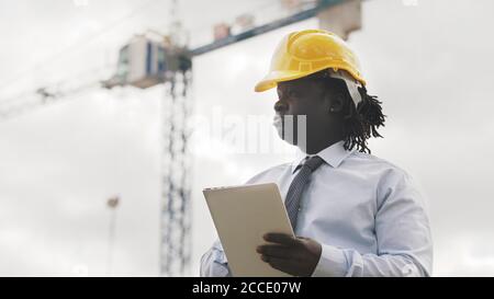 Ritratto di un uomo africano con hardhat utilizzando la tavoletta sul cantiere e osservando il lavoro. Foto di alta qualità Foto Stock