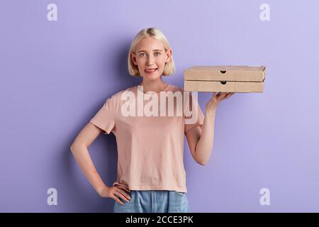 donna allegra affamata che mostra la sua lingua, la lingua di messa fuori, tenendo le scatole della pizza, guardando la macchina fotografica. primo piano ritratto, isolato violetto backgroun Foto Stock