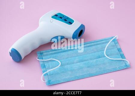 Maschera medica e termometro elettronico a infrarossi per la misurazione della temperatura, sfondo rosa. Pandemia di coronavirus (COVID-19). Concetto di prevenzione dell'influenza Foto Stock
