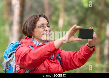 Una donna anziana con uno zaino nella foresta scatta una foto utilizzando il suo smartphone. Concetto di stile di vita attivo all'età dopo il 60. Foto Stock