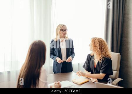 Gruppo di donne d'affari che si incontrano in ufficio. Donna bionda vestita in tuta formale che condivide le sue idee con due colleghi seduti alla scrivania. Foto Stock