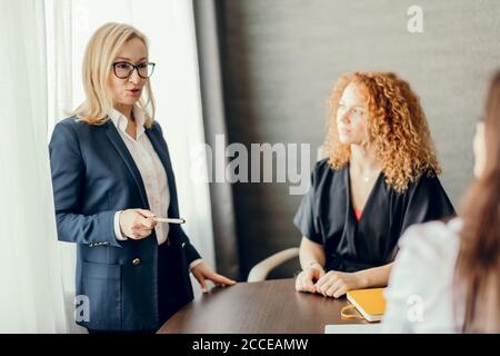 Esperti di marketing femminile seduti intorno al tavolo e guardando la donna speaker in riunione informale. Bionda dirigente caucasico femminile che dà un presentatio Foto Stock