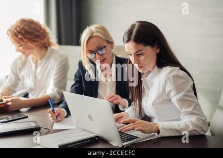 Tre stilisti femminili di moda si incontrano negli interni moderni degli uffici, discutendo di business plan e idee utilizzando un computer portatile. Foto Stock