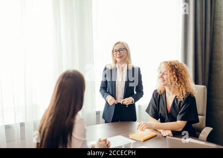 Esperti di marketing femminile seduti intorno al tavolo e guardando la donna speaker in riunione informale. Bionda dirigente caucasico femminile che dà un presentatio Foto Stock