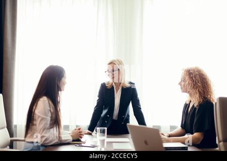 Le colleghe discutono insieme dei piani di lavoro, seduti al tavolo dell'ufficio. Il intern femminile dai capelli rossi pone domande al capo del reparto biondo. Capo Foto Stock