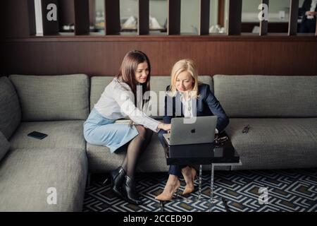 Due donne sorridenti che lavorano in linea con un computer portatile seduto sul divano in ufficio. Foto Stock