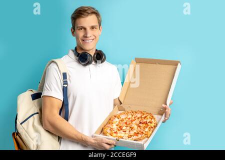 ritratto di giovane amante della pizza caucasica isolato su sfondo blu, maschio in t-shirt bianca con le cuffie tenere la pizza nelle mani, gustare mangiare, delive Foto Stock