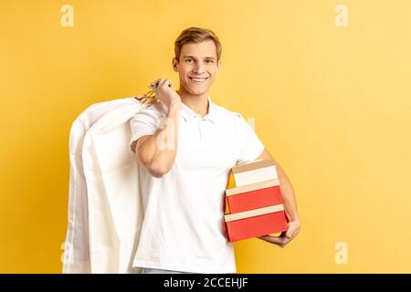 Consegna, concetto di corriere. Ritratto di sorridente giovane uomo caucasico consegna in t-shirt con vestiti di imballaggio sul retro e regali in mani per il cliente Foto Stock