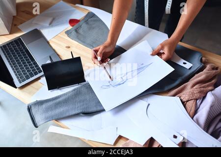Vista dall'alto su come creare uno schizzo per una nuova collezione di moda. Laptop, carte, tessuti su tavolo Foto Stock