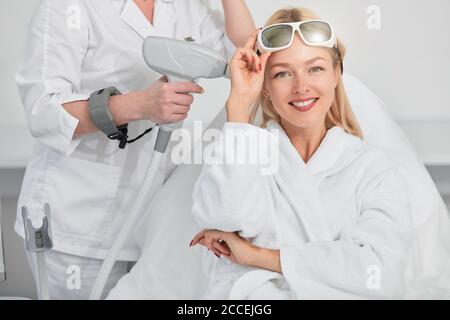 allegra donna bionda sorridente con gli occhiali sulla fronte che si prepara per le procedure laser della pelle al salone di bellezza. Cura di bellezza. Alta risoluzione. Si verifica Foto Stock