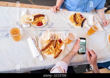 Uomo e donna si siedono sulla tavola della birra con piatti pieni di salsiccia e patatine rosse e bianche, con birra da tazze di plastica Foto Stock
