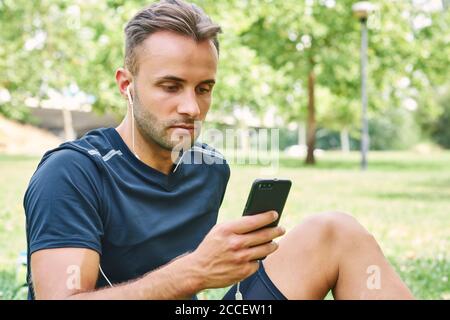 Uomo con il suo smartphone seduto sull'erba in un parco dopo l'allenamento. Vita sana e sana. Concetto di salute e sport. Foto Stock