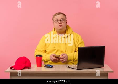 Giovane uomo con cappuccio giallo seduto dietro il monitor del computer portatile guarda la telecamera in disillusione mentre si guarda in movimento durante il suo turno di lavoro Foto Stock