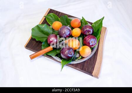 Frutta estiva, prugne e albicocche, con foglie di prugne e albicocca in un piatto d'argento su un tagliere di legno con coltello da affettare, isolato su una panca bianca Foto Stock