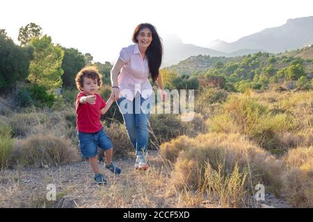 Madre e figlio che giocano facendo jogging sul campo su un giorno fuori tenendo le mani e ridendo Foto Stock