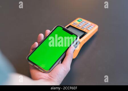 vista a spalla di un uomo che tiene lo smartphone con schermo verde sul terminale pos sul contatore. sfocatura dello sfondo Foto Stock