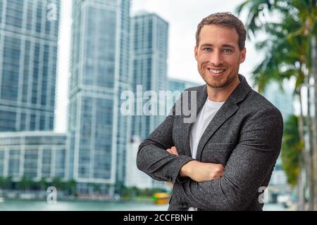 Felice sorridente giovane uomo d'affari caucasico fiducioso con le braccia incrociate ritratto indossando un blazer professionale casual elegante su sfondo urbano della città Foto Stock
