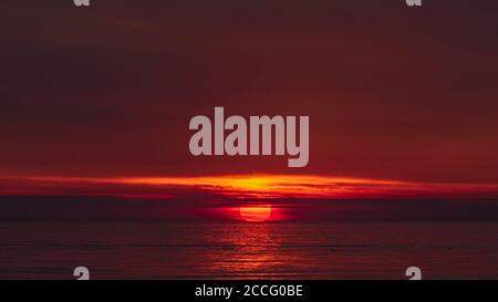 il momento del tramonto quando il sole tocca l'orizzonte, camuffandosi dietro nuvole sottili e colorando il rosso dell'acqua Foto Stock