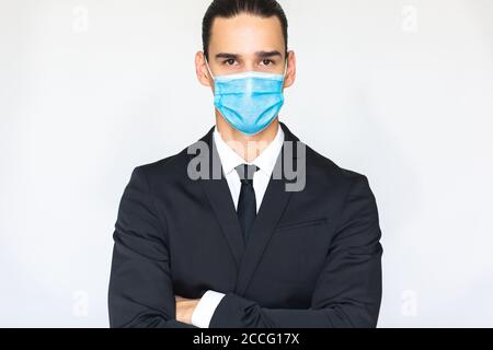 Giovane uomo d'affari sicuro in tuta indossando una maschera medica e guardando la macchina fotografica con le braccia incrociate. Ritratto isolato su sfondo bianco. Foto Stock