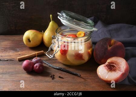 Conservando pesche, pere e prugne con cannella e chiodi di garofano in un vaso di vetro su legno rustico per avere frutta in scatola in inverno, vintage scuro e moody stile, Foto Stock