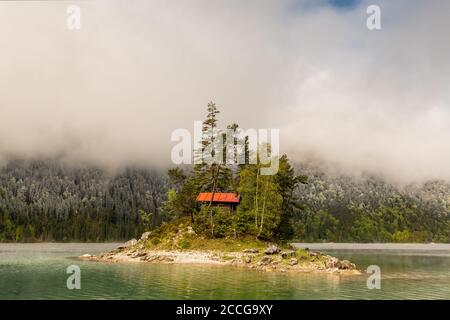 Piccola isola solitaria con cabina in legno e tetto rosso, nel bel mezzo dell'Eibsee sotto lo Zugspitze nelle Alpi bavaresi del Wetterstein Foto Stock