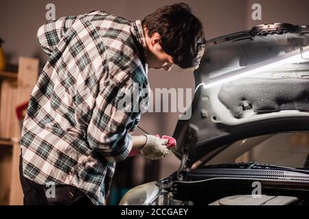 un giovane meccanico maschile in una camicia e guanti controlla il livello dell'olio motore in una vettura nera Foto Stock
