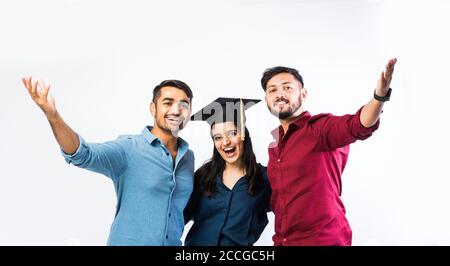 Bella giovane studentessa indiana laureata che celebra il successo con il maschio Amico mentre indossa il cappello della cerimonia di laurea su sfondo bianco Foto Stock