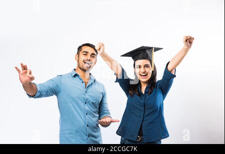 Bella giovane studentessa indiana laureata che celebra il successo con il maschio Amico mentre indossa il cappello della cerimonia di laurea su sfondo bianco Foto Stock