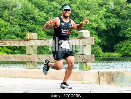 Babylon, New York, USA - 12 agosto 2018: Un corridore che corre un sentiero 10K dà due segnali di pace mentre sta attraversando un ponte su un lago. Foto Stock