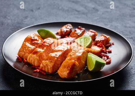 primo piano di filetti di salmone teriyaki appiccicosi cotti al forno servito su un piatto nero con i cunei di lime su un tavolo in cemento Foto Stock