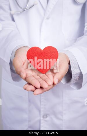 Il giovane medico asiatico regge un cuore rosso con due mani. Vesti con uniforme di abito medico con stetoscopio in un ospedale. Foto Stock
