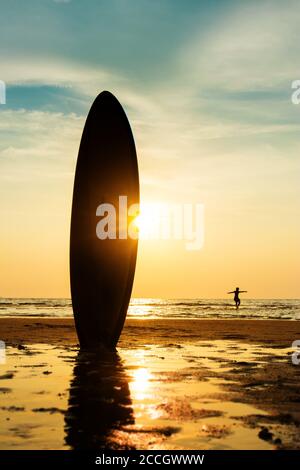 Silhouette di surf uomo con una tavola da surf sulla spiaggia di mare al tramonto. Bel modello di uomo asiatico nei suoi anni 20. Foto Stock