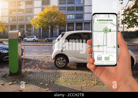Con un telefono cellulare sullo sfondo di auto elettrica per il car sharing in carica Foto Stock