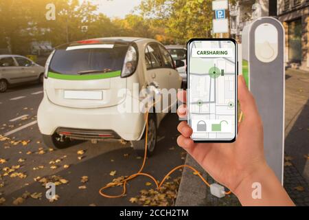 Con un telefono cellulare sullo sfondo di auto elettrica per il car sharing in carica Foto Stock